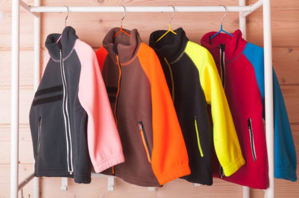 Colorful fleece jackets are hanging on hangers near wooden wall in a rack